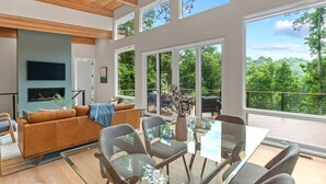 A spacious living area with a glass table and chairs, a brown sofa, and a large window offering a view of a lush green landscape. - Mtn View, 3 Miles to DT AVL, King Bed, Fire Pit (Asheville)
