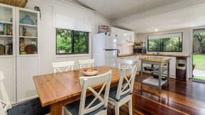 A modern kitchen with white cabinetry, a wooden dining table, and a view of the garden through large windows. - Nelly's Hidden Cottage (BONGAREE)