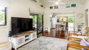 A spacious living area with a flat-screen TV on a wooden stand, a dining table with chairs, and a kitchen in the background. - Nelly's Hidden Cottage (BONGAREE)