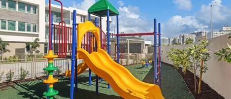 A playground with a yellow slide, climbing structures, and a green-roofed playhouse.
