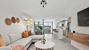 A modern living room with a sofa, coffee table, and decorative items. There is a kitchen area with a refrigerator and a dining table. - Poolfront by Isla Verde Beach (Carolina)