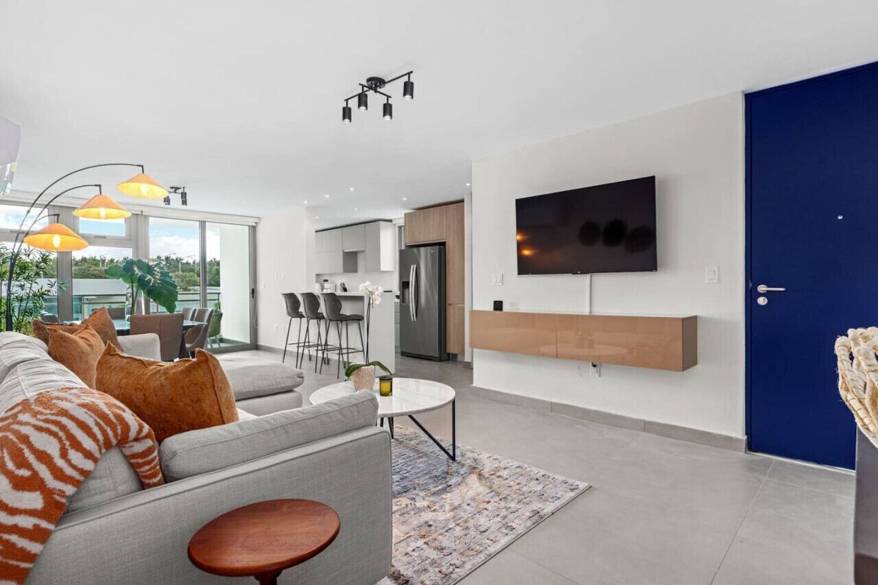 A modern living room with a grey sofa, orange and white cushions, a round coffee table, and a TV mounted on the wall.
