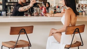 Eine Bar mit zwei Holzsesseln, einer Theke mit verschiedenen Flaschen und einer Person, die ein Getränk ausschenkt.