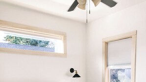 Een net opgemaakt bed met witte lakens en een bloemenkussen, een nachtkastje, een wandlamp en een raam met zonwering.