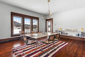 A dining area with a wooden table, chairs, a patterned rug, and a bookshelf with books and decorative items. - Elegant Country Escape – Unique Home with Timeless Charm and Style (Margaretville)