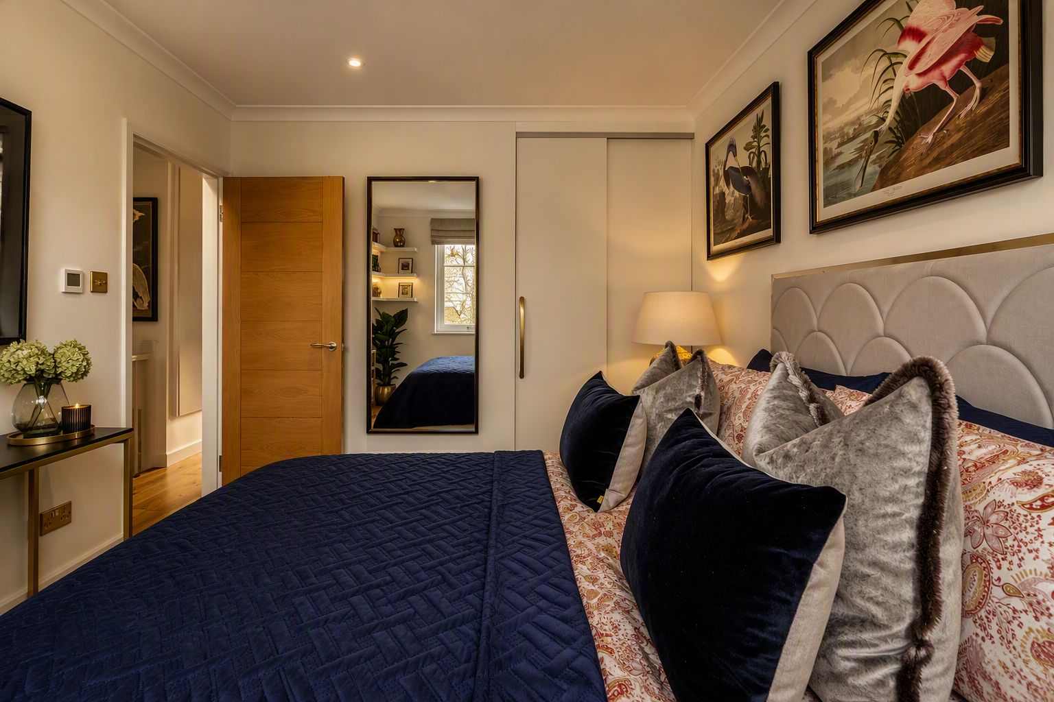 A bedroom with a large bed, a bedside table with a lamp, a mirror, and a framed picture on the wall.