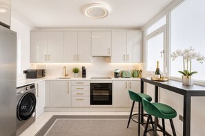 A modern kitchen with white cabinets, a built-in oven, a washing machine, and a bar area with green chairs.