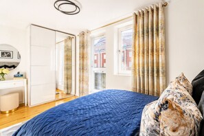 Une chambre à coucher comprenant un lit, un bureau, une chaise, un miroir et une vue sur les bâtiments par la fenêtre.