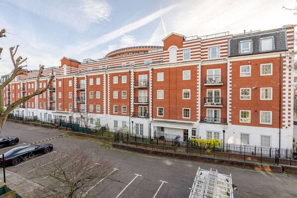 A multi-story residential building with a brick facade, multiple balconies, and a parking area in front.
