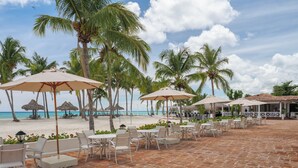 Un patio frente a la playa con mobiliario exterior y sombrillas, palmeras y vista al océano.
