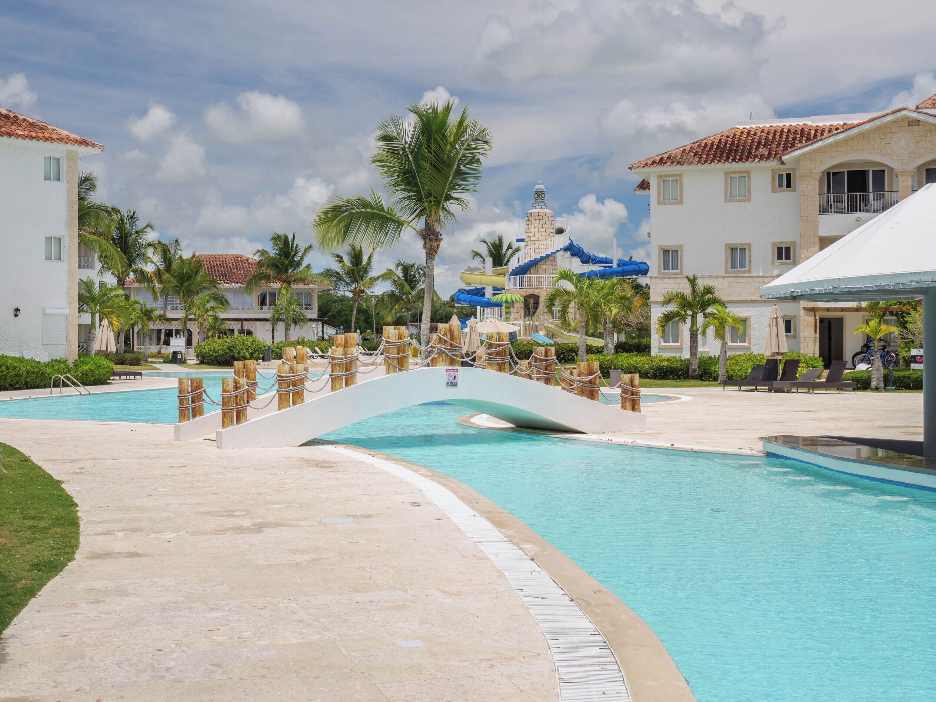 A resort pool area with a bridge, palm trees, and buildings with tiled roofs.