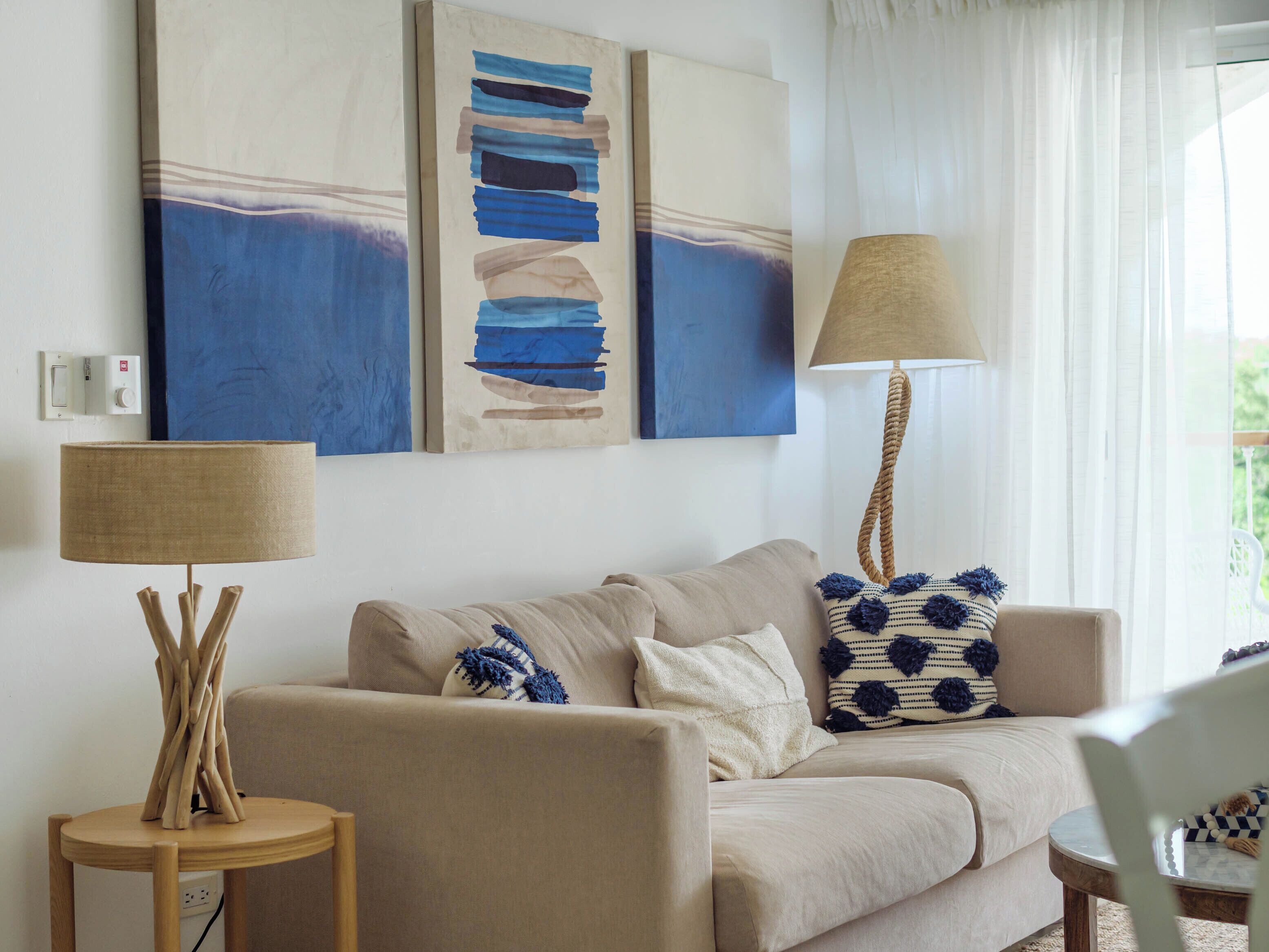 Un salon avec un canapé beige, une petite table d’appoint ronde en bois sur laquelle se trouve une lampe, et deux tableaux abstraits accrochés au mur.