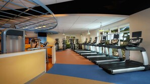 Ein Fitnessstudio mit Laufbändern, Trainingsgeräten und einem großen Fenster mit Blick auf Palmen.