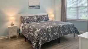 A bedroom with a bed, two bedside tables, a nightstand, a window with blinds, and a painting on the wall. - Willow Bend Condo | Close to Barefoot Golf Courses (North Myrtle Beach)