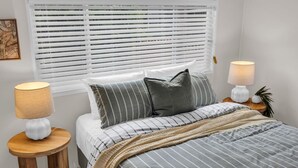 A bedroom with a bed, striped bedding, two bedside lamps, a window with blinds, and a small round table with a lamp. - Settle Inn: Broadwater, Pet Friendly (Broadwater)