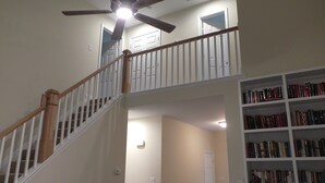 A multi-level interior with a wooden staircase, a ceiling fan, and a bookshelf filled with books.