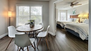 Una habitación con una mesa redonda de comedor, sillas blancas, un ventilador de techo, una cama con cabecera de madera y una ventana con persianas.