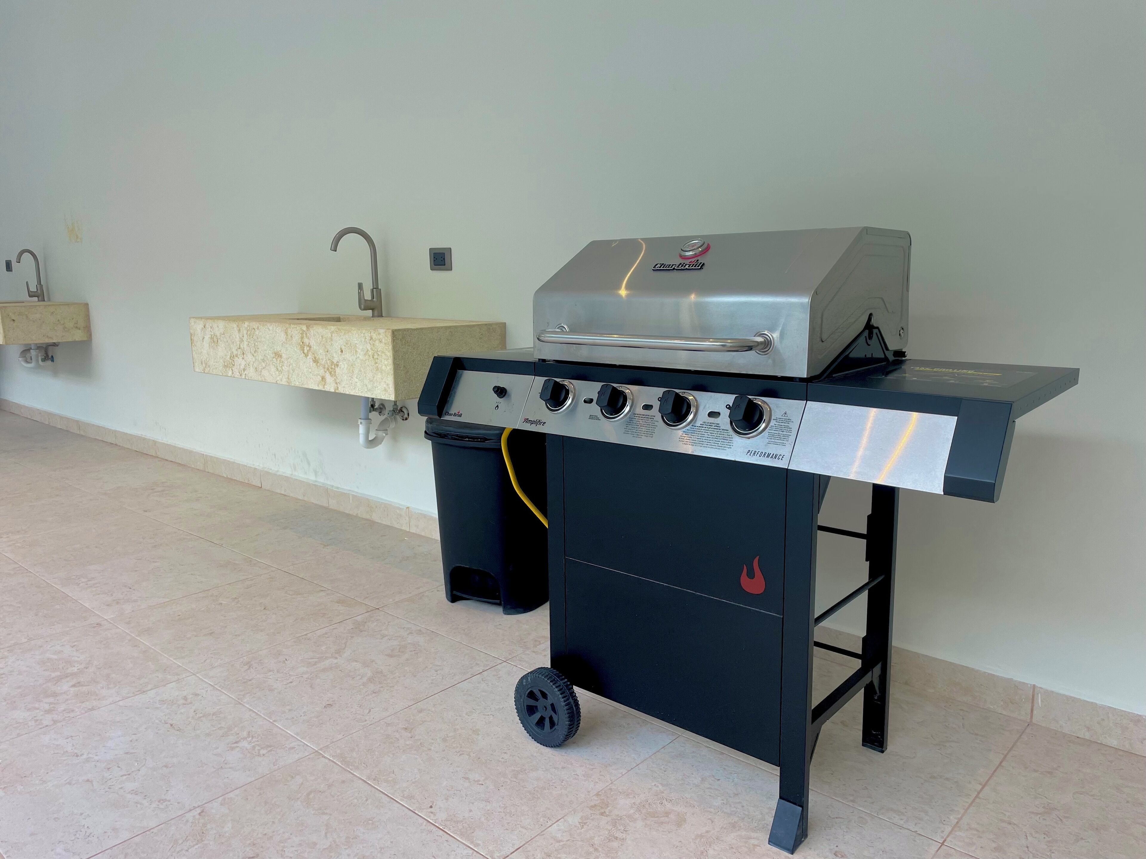 Een draagbare gasgrill met vier branders en een bijzettafeltje, geplaatst in een ruimte met betegelde vloer en een aan de muur bevestigd spoelbakje.