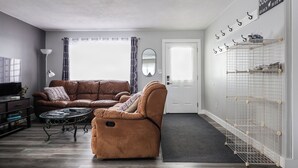 A living room with a brown sofa, a glass coffee table, and a TV on a stand. There is a door with a coat rack and a white shelf with items on it. - Le Lodge - Spacious Home (Cochrane)