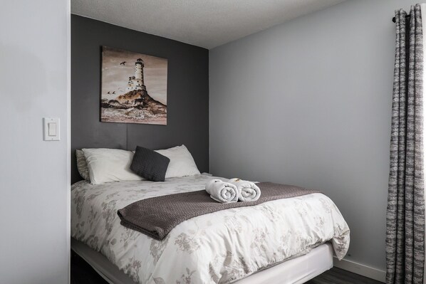 Un lit bien fait, recouvert d’une couette à motifs floraux, d’un plaid gris et de deux serviettes enroulées. Un tableau représentant un phare est accroché au mur au-dessus du lit.