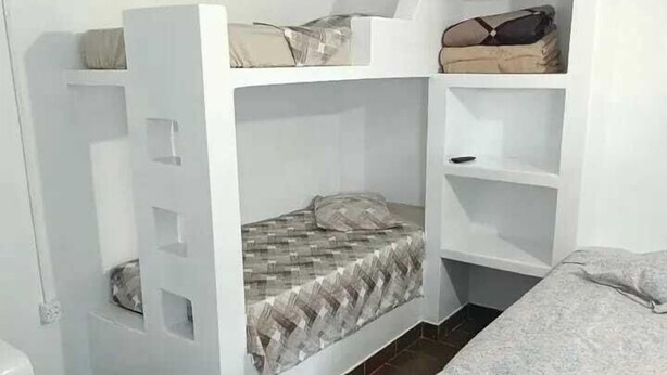 A compact room with a bunk bed, a small shelf unit, and a bed with a patterned bedspread. - Pousada Tia Carmem (Ilha Comprida)