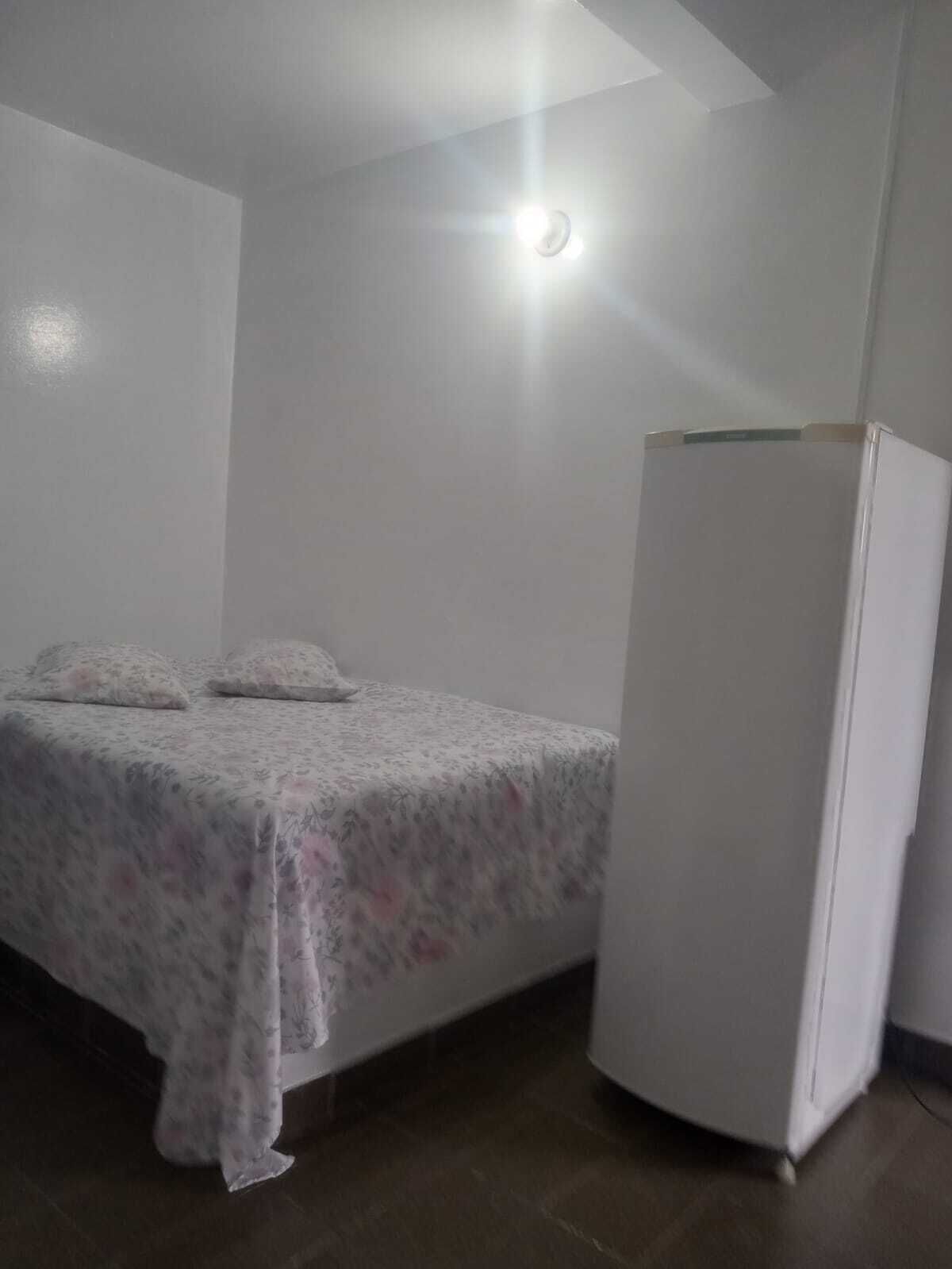 A small, sparsely furnished room with a bed, a white refrigerator, and a ceiling light.