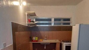 A compact kitchen with a dining table covered by a red tablecloth, wooden chairs, a built-in oven, and a small countertop with a sink. - Pousada Tia Carmem (Ilha Comprida)