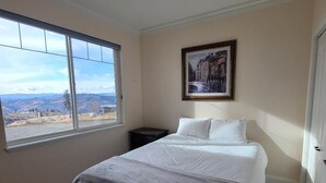 A bedroom with a large window, a bed with white bedding, a dark nightstand, and a framed picture on the wall.