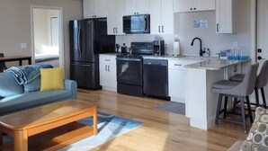 A modern kitchen with white cabinets, a black refrigerator, a microwave, and a dishwasher. There is a wooden coffee table and a gray sofa with yellow cushions.