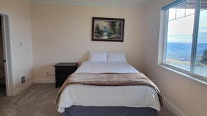 A bedroom with a bed, a nightstand, a painting, and a window with a view.