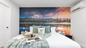 Une chambre avec une grande fresque murale représentant une montagne et un lac, une couverture bleue moelleuse, un livre et une tasse à café posés sur le lit.