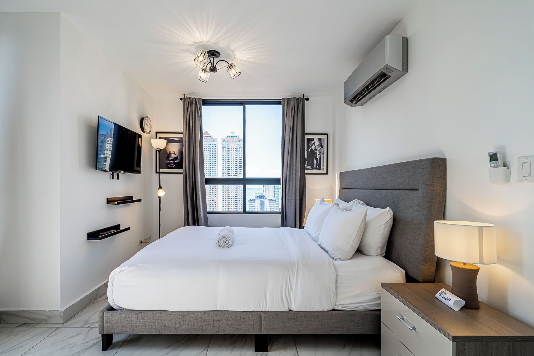 Ein modernes Hotelzimmer mit einem großen Bett, einem Flachbildfernseher, Blick auf die Stadt und einer Deckenleuchte.