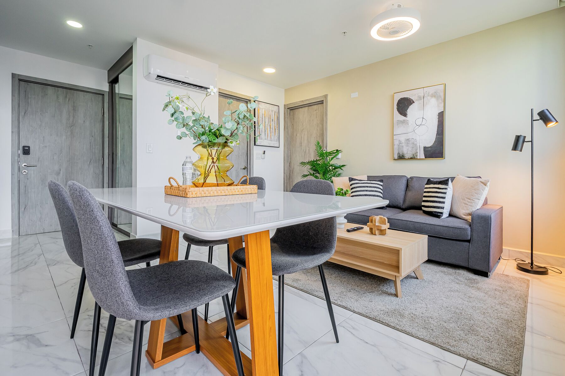 A modern living room with a dining table, grey upholstered chairs, a grey sofa with striped cushions, a wooden coffee table, and a floor lamp.
