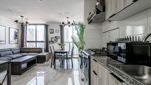 Ett modernt kök med en inbyggd spis, en matplats med en mörkfärgad soffa och ett litet bord, samt ett stort fönster med utsikt över höga byggnader.