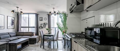 Una cocina moderna con estufa integrada, un comedor con sofá oscuro y una mesita, y un ventanal con vistas a edificios altos.