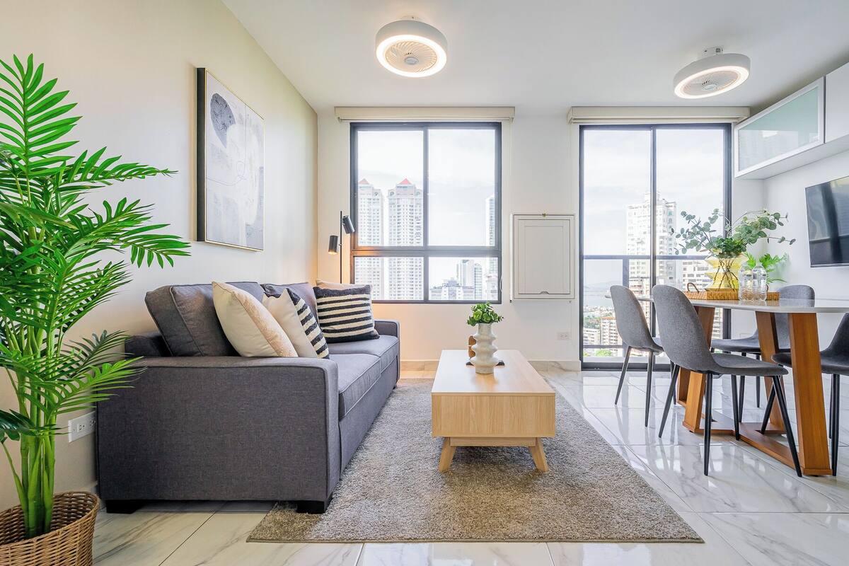 Ein modernes Wohnzimmer mit grauem Sofa, einem Holztisch und einem Essbereich mit Tisch und Stühlen. Große Fenster bieten einen Blick auf die Stadtlandschaft.