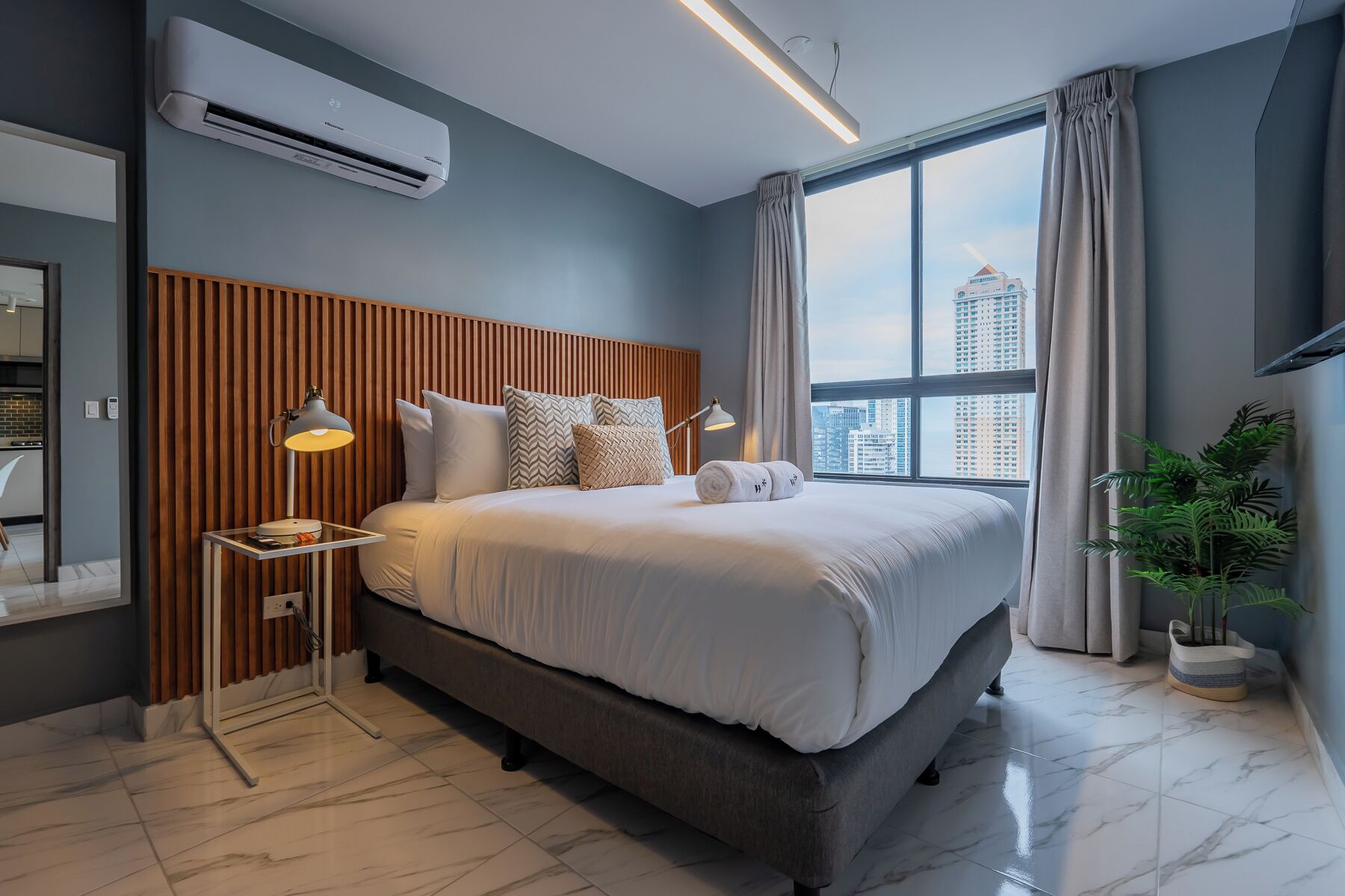 Moderni hotellihuone, jossa on suuri sänky, puinen pääty, näkymä kaupunkiin ja ruukussa oleva kasvi.