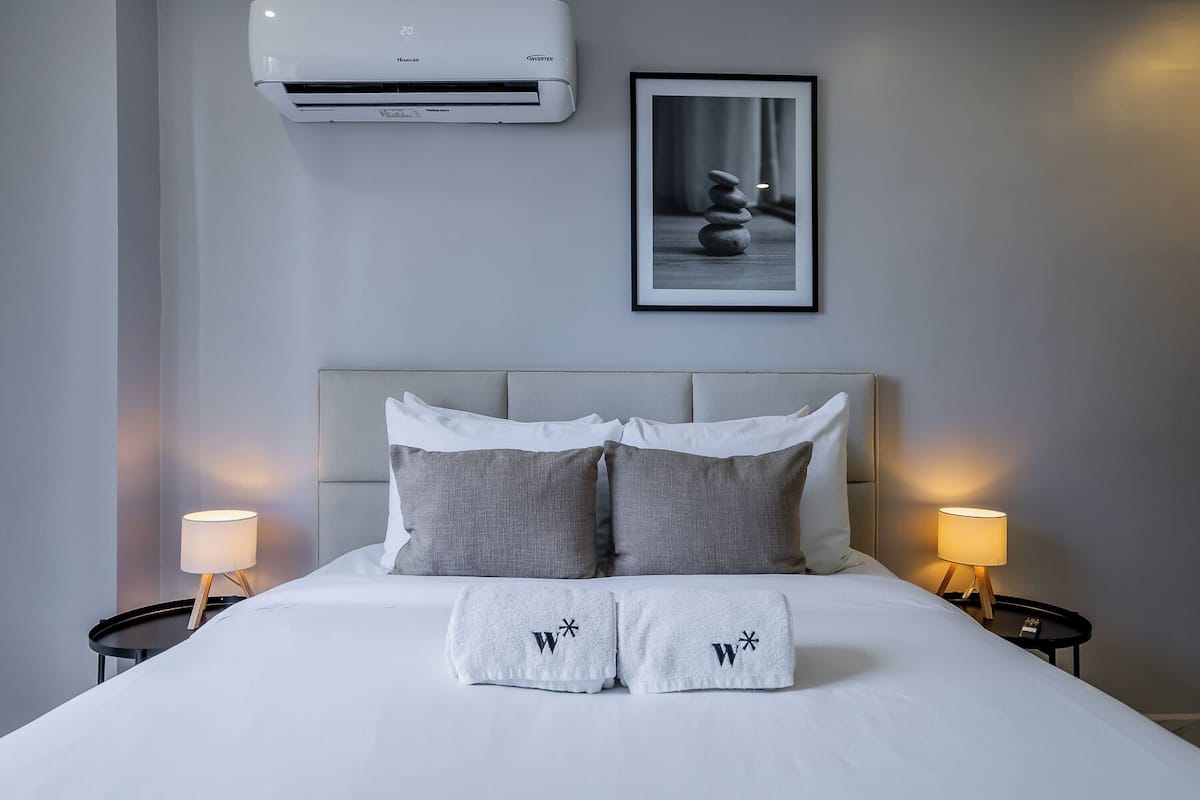 Ein Hotelzimmer mit einem Bett, zwei Nachttischlampen, einem gerahmten Bild an der Wand und einer Klimaanlage über dem Bett.