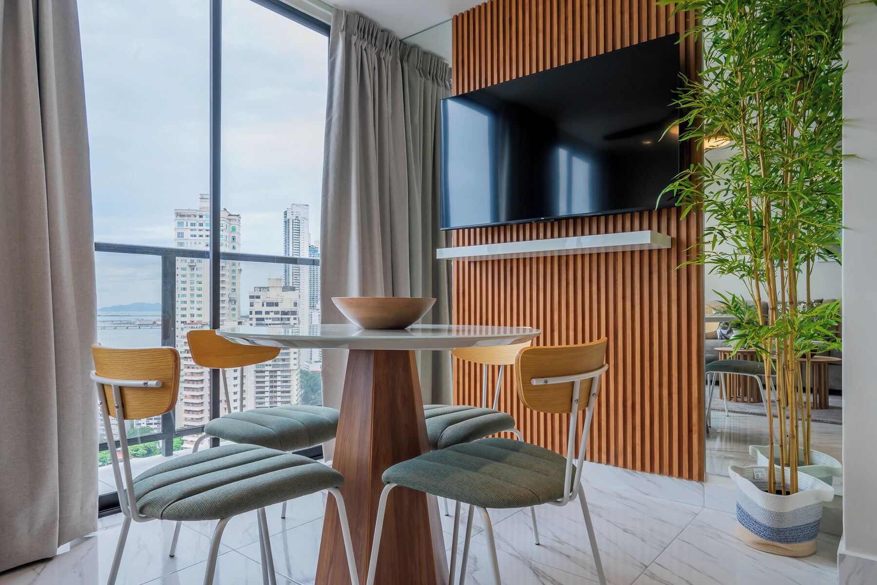 Un salon moderne comprenant une table à manger, des chaises, un téléviseur et une grande fenêtre donnant sur la ville.