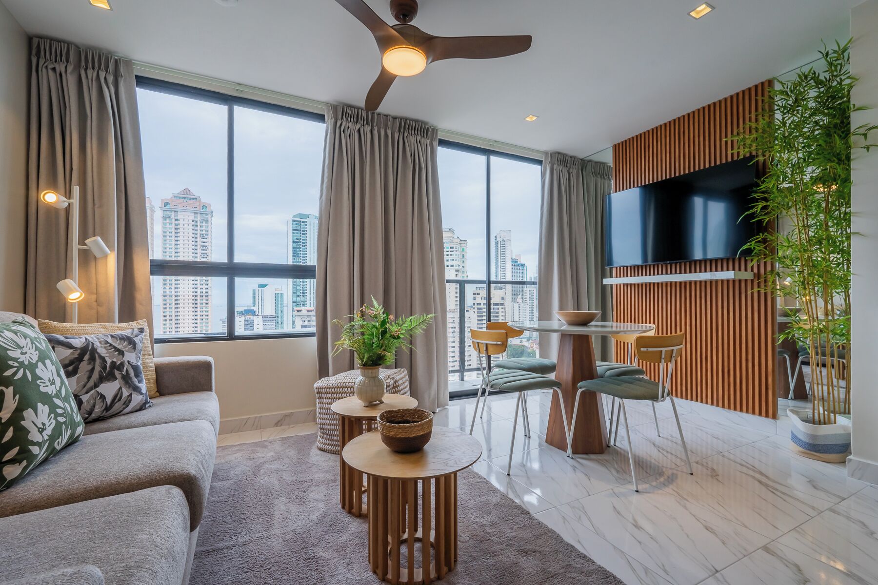 Un salon moderne comprenant un canapé, une table basse, un coin repas avec des chaises, un téléviseur fixé sur un panneau en bois et une vue sur la ville à travers de grandes fenêtres.