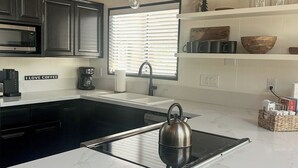 A modern kitchen with dark cabinets, a white countertop, a kettle, and a window with blinds.