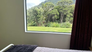 A bedroom with a bed, a window with a view of a forest and a hill, and a curtain. - Remote Pelorus Sound Kiwi Bach on the Beach with Great Views (Totaranui)