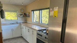 A kitchen with white cabinets, a stainless steel refrigerator, a stove, and a window with a view of greenery. - Remote Pelorus Sound Kiwi Bach on the Beach with Great Views (Totaranui)