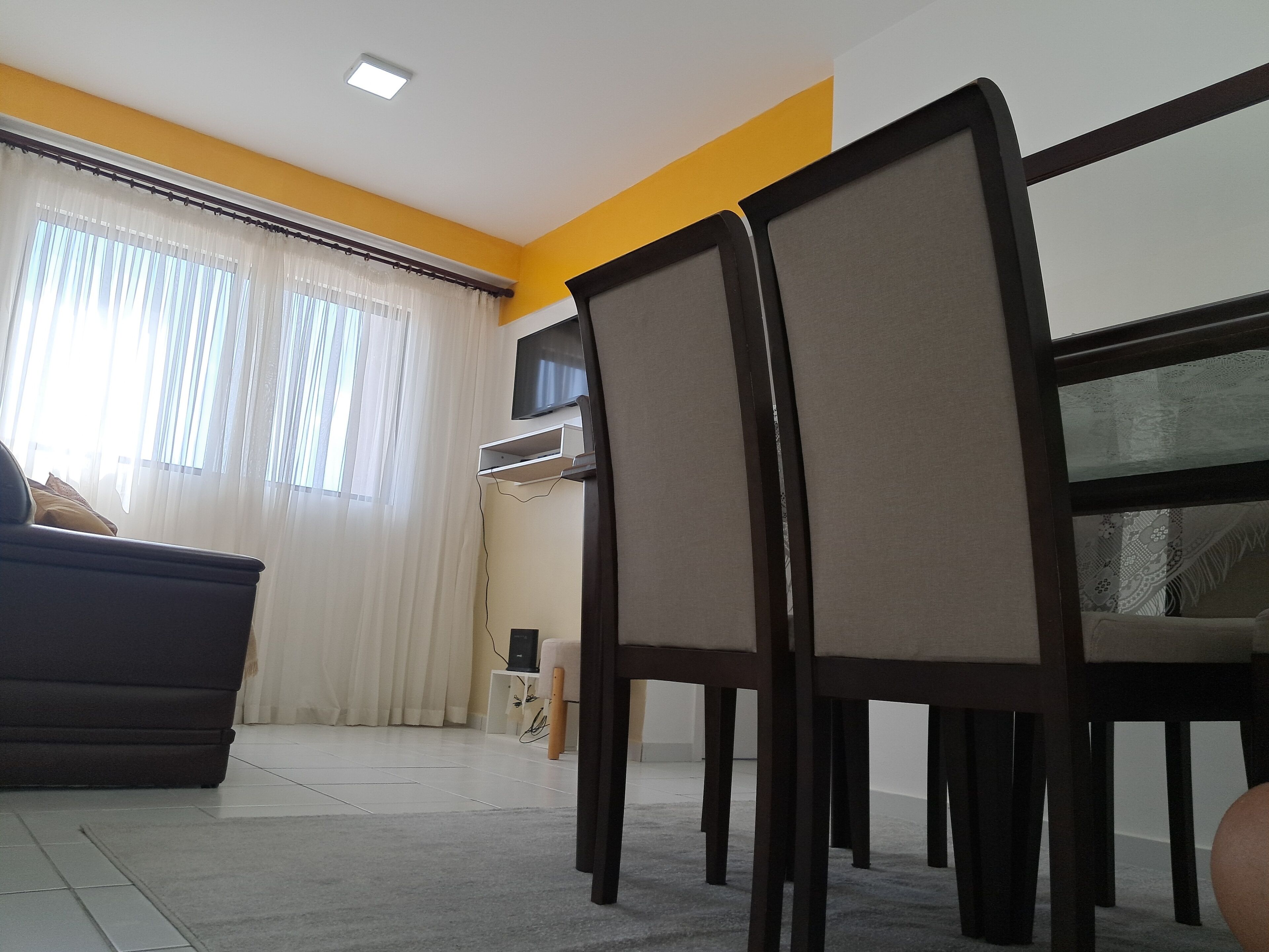 A living room with a sofa, a dining table with chairs, a TV mounted on the wall, and a window with curtains.