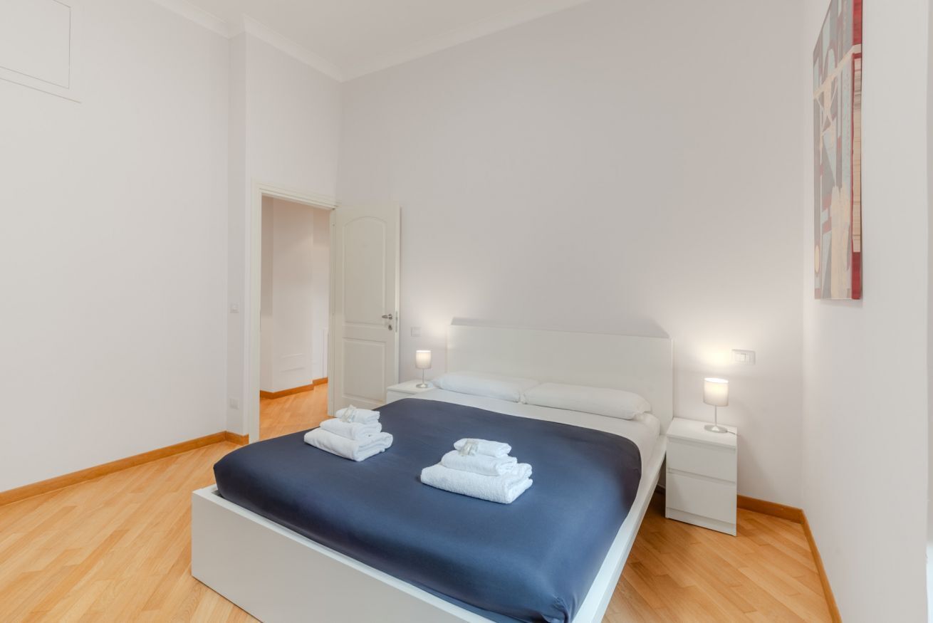Una habitación de hotel limpia y moderna con una cama bien hecha, mesitas de noche con lámparas y una puerta que da a otra habitación.