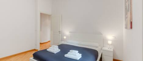 Una habitación de hotel limpia y moderna con una cama bien hecha, mesitas de noche con lámparas y una puerta que da a otra habitación.
