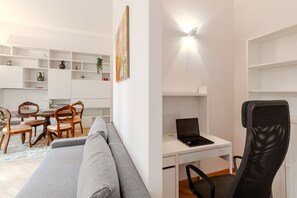 Una sala moderna con un sofá gris, una mesa de comedor de madera y sillas, un escritorio blanco con una computadora portátil, y una silla de malla negra.