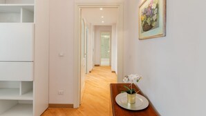 A hallway with a wooden floor, white walls, and a framed painting. There is a small table with a vase of flowers. - Contemporary Flat at Trastevere (Roma)