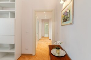 A hallway with a wooden floor, white walls, and a framed painting. There is a small table with a vase of flowers. - Contemporary Flat at Trastevere (Roma)