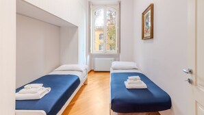 A narrow hotel room with two beds, a window with a view of buildings, and a framed picture on the wall. - Contemporary Flat at Trastevere (Roma)